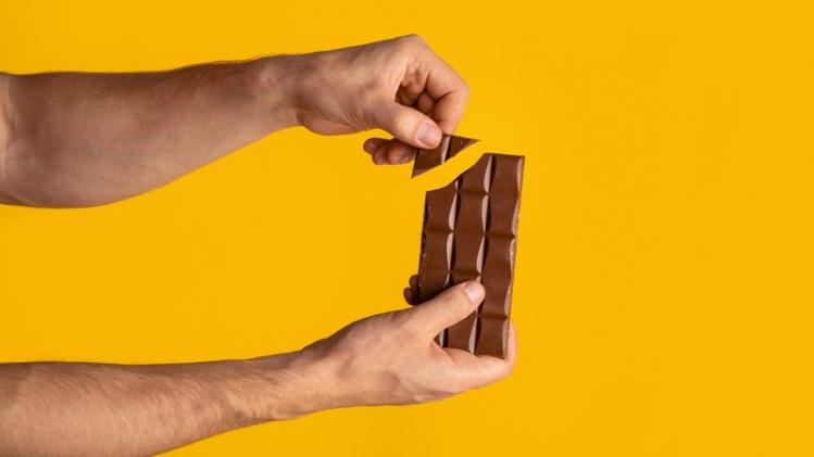 Closeup of man breaking piece of chocolate from bar, orange background. Copy space. Panorama